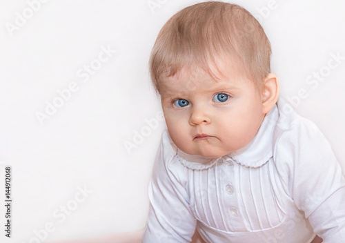 Beautiful girl with blue eyes in white dress stands on all fours on the floor and seriously looking at the camera. Portrait of a baby in 9 months