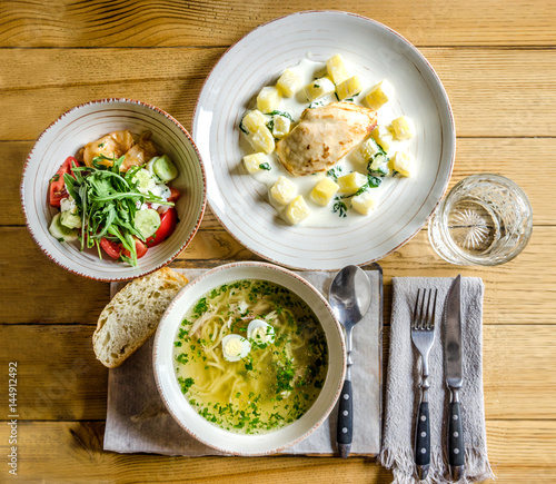 Dinner: soup with vermicelli and egg, chicken wing with pineapple, salad with vegetables and herbs, bread, a glass of water and cutlery, on a wooden table. View from above photo