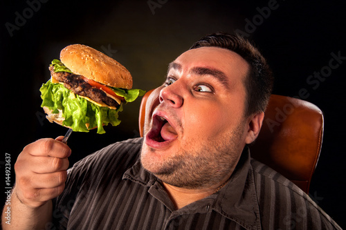 Diet failure of fat man eating fast food hamberger. Happy overweight person with wide-open mouth who spoiled healthy food by greedily eating huge hamburger on fork. Junk meal leads to obesity.