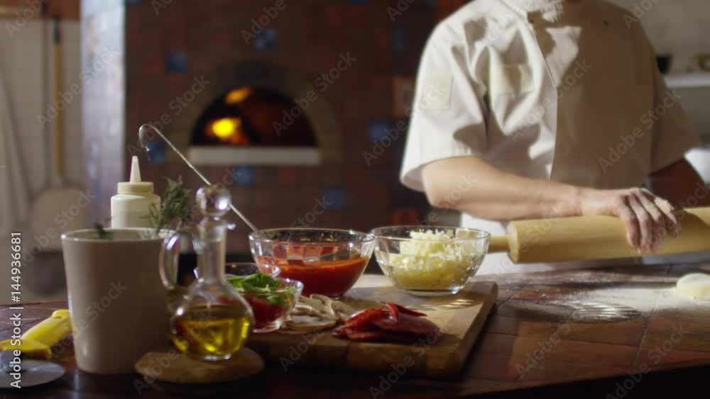 Tracking shot in slow motion of baker rolling dough on table in restaurant kitchen. There are bowls with pizza toppings on it Tracking shot in slow motion of baker rolling dough on table in restaurant kitchen. There are bowls with pizza toppings on it