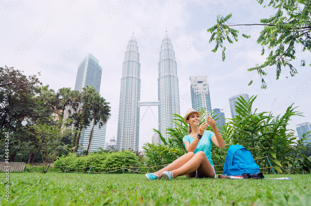 Naklejka premium Koncepcja podróży. Dość młoda kobieta w kapeluszu za pomocą smartfona siedząc na trawie przed Petrones Twins Tower w Kuala-Lumpur.