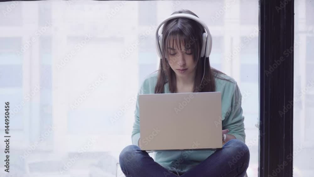 Pensive brunette woman in headphones is sitting against the window with a laptop on her knees and writing something Pensive brunette woman in headphones is sitting against the window with a laptop on her knees and writing something