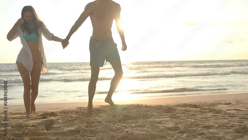 Young happy couple walking on the beach and holding hands of each other at sunset. Man and woman going on sea shore during beautiful sunrise. Slow motion Young happy couple walking on the beach and holding hands of each other at sunset. Man and woman going on sea shore during beautiful sunrise. Slow motion