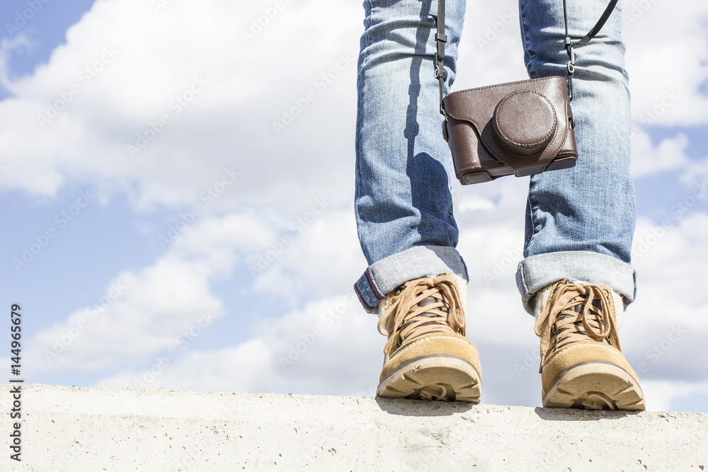 Young woman with feet clad in boots, standing on a concrete wall, holding a camera in her hands. Against the background of the blue sky and white clouds. Young woman with feet clad in boots, standing on a concrete wall, holding a camera in her hands. Against the background of the blue sky and white clouds.