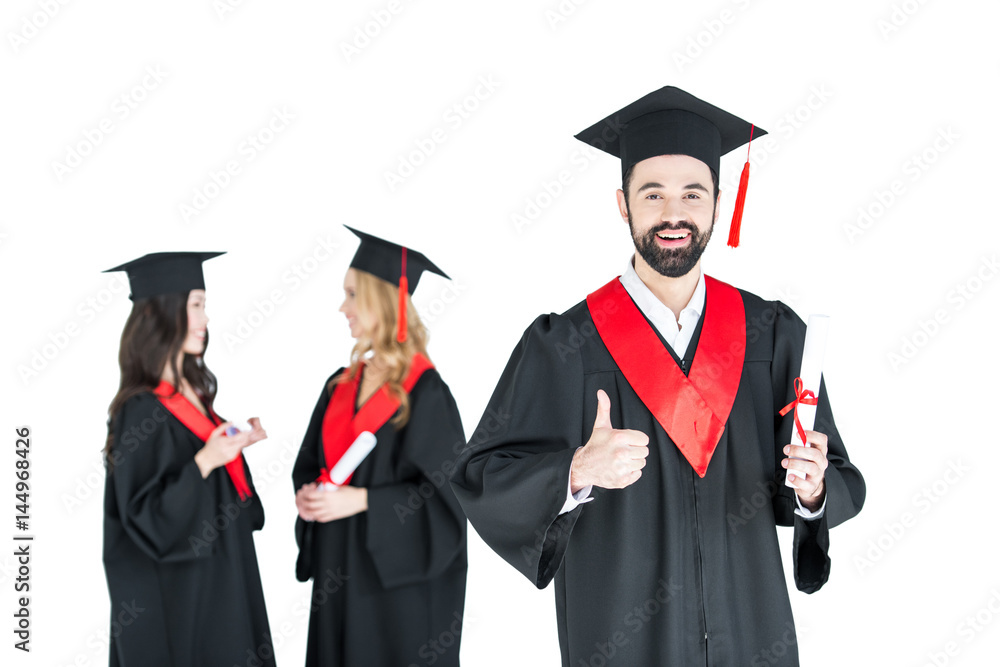 Happy young man in academic cap holding diploma and showing thumb up
