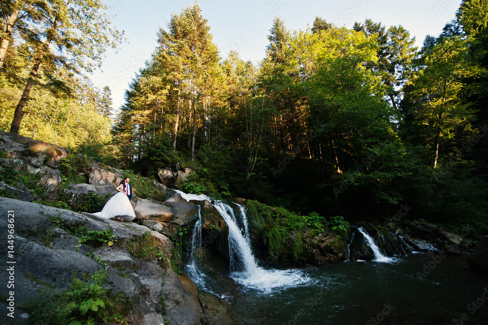 Lovely wedding couple against waterfall on sunset at Carpathian mountains.