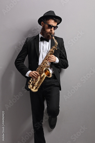 Jazzer playing on a saxophone and leaning against gray wall