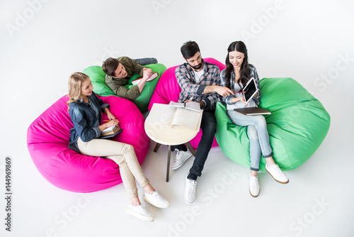 students sitting on beanbag chairs and studying with digital tablet in studio on white