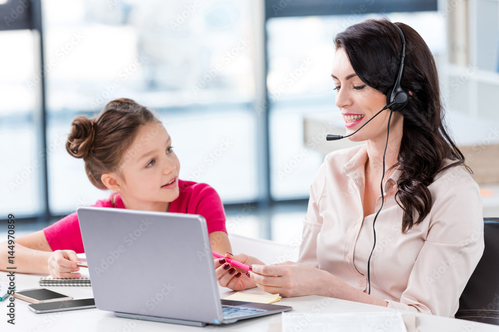 Smiling little girl looking at happy mother in headset working with laptop Smiling little girl looking at happy mother in headset working with laptop