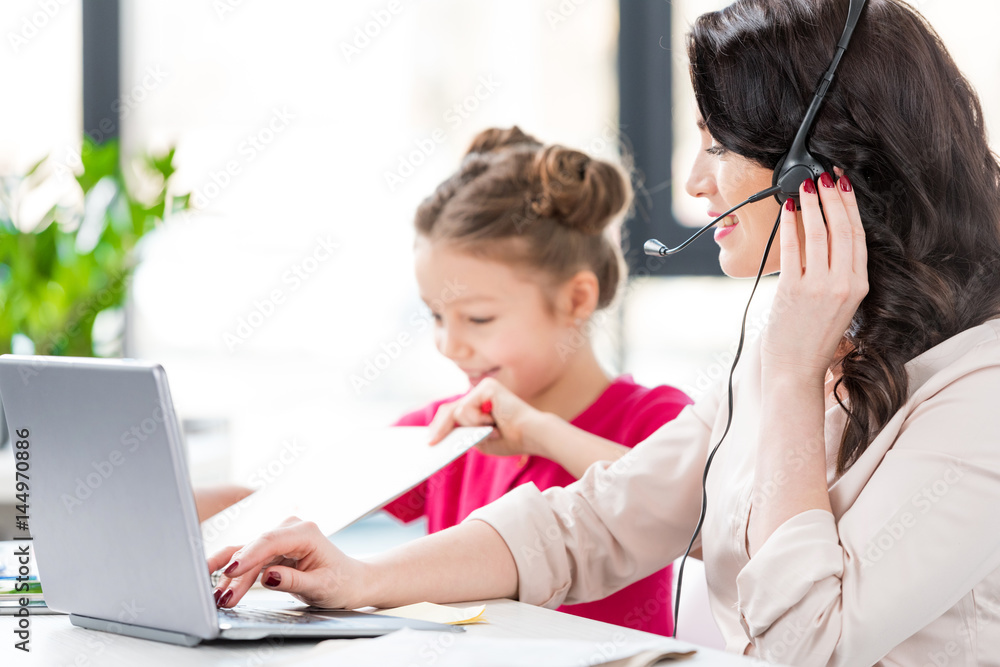 Cute little girl sitting near smiling mother in headset working with laptop