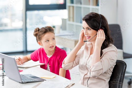 Cute little girl looking at happy mother in headset working with laptop