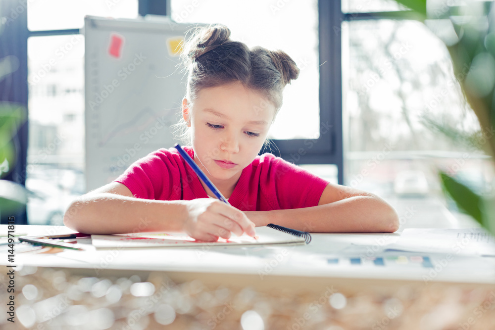 Concentrated little girl sitting at desk and drawing in office