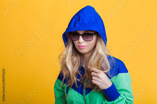 portrait of beautiful blond woman in sunglasses and blue green hooded jacket on yellow background. hipster summer. © producer