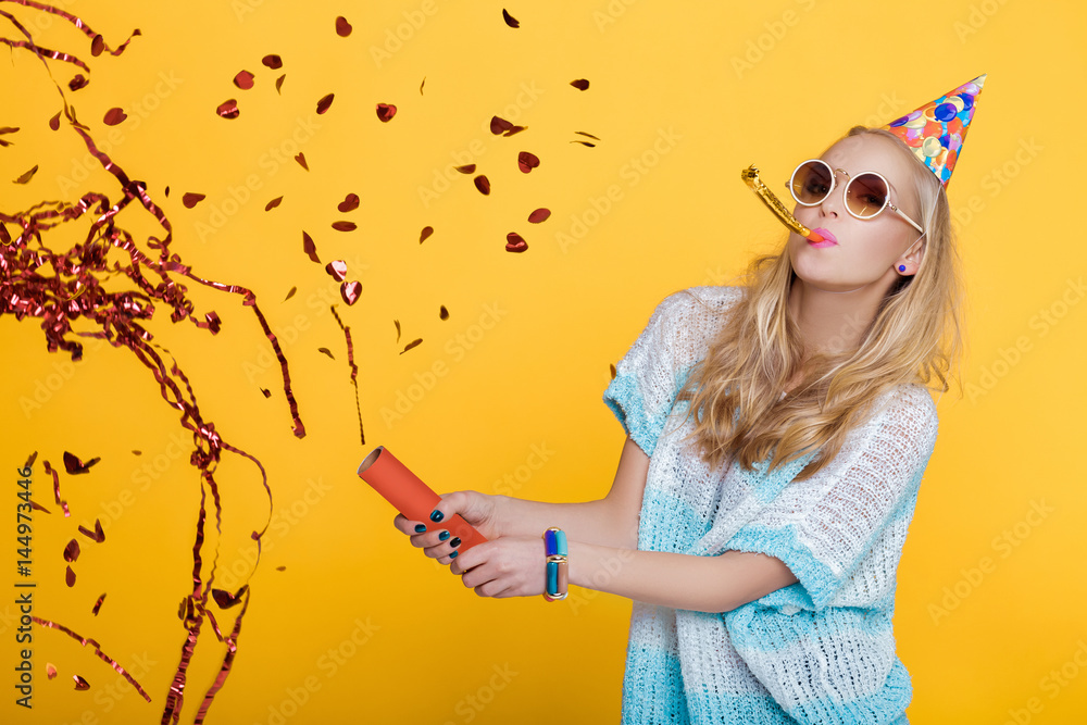 portrait of funny blond woman in birthday hat and red confetti on yellow background. Celebration and party. - obrazy, fototapety, plakaty 