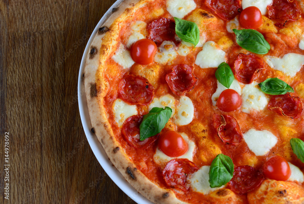 Top view of neapolitan pizza with pepperoni, mozzarella and cherry tomatoes on a white plate for a dinner in italian restaurant. Italy food. Pizza from wooden oven. Close up. Macro. Top view of neapolitan pizza with pepperoni, mozzarella and cherry tomatoes on a white plate for a dinner in italian restaurant. Italy food. Pizza from wooden oven. Close up. Macro.