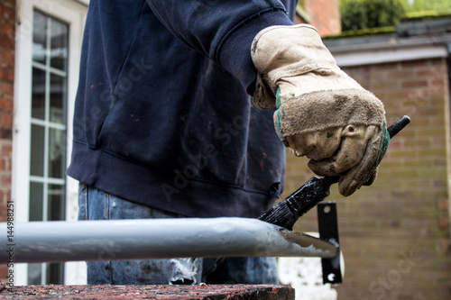 Painter decorator metalworker paints metal iron railing black with baggy jumper and workman’s gloves in back garden of residential job - BIY craftsman