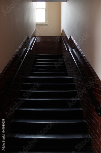 Light hits dark stairwell of old nineteenth century school house in England with hanging light  burgundy red tiles and shiny black painted stairs