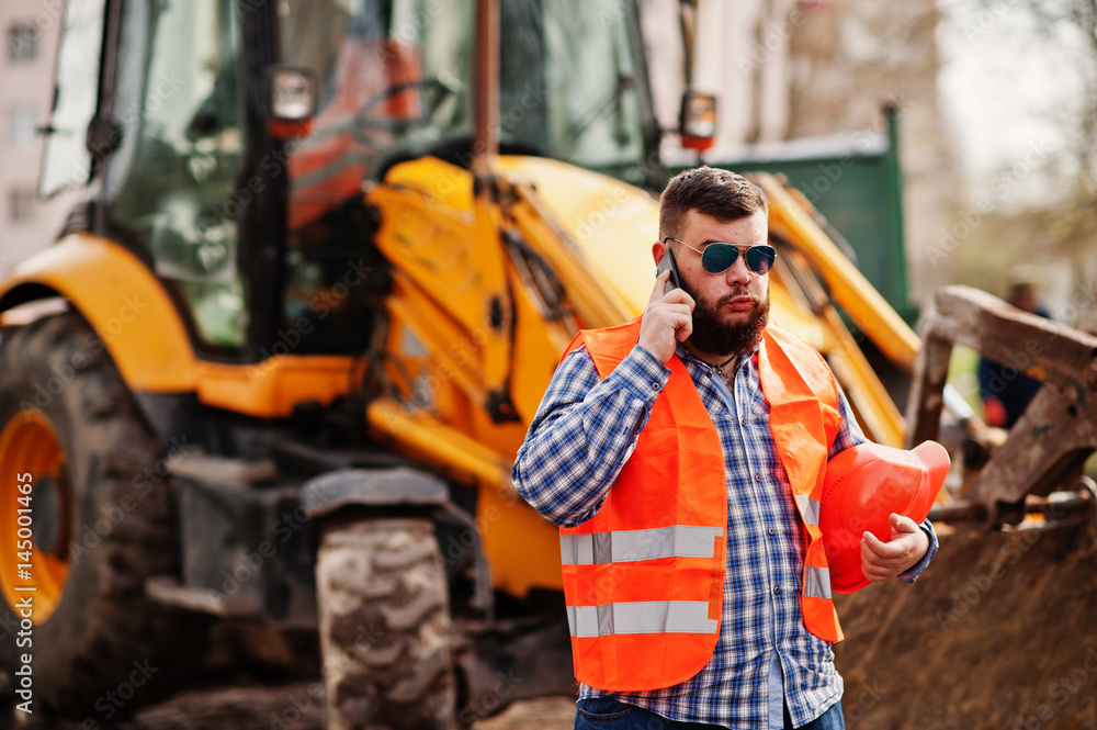 Obraz premium Brutal beard worker man suit construction worker in safety orange helmet, sunglasses against traktor with mobile phone at hand.
