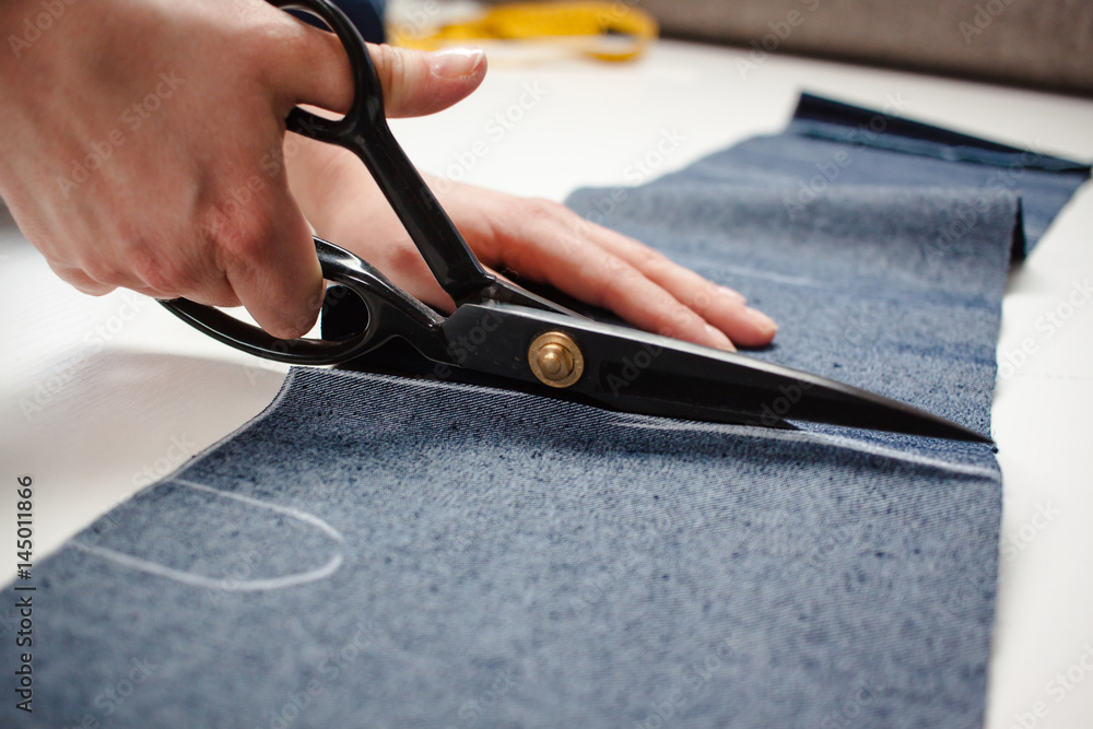 Hands of seamstress cutting a jeans fabric with scissors on white table