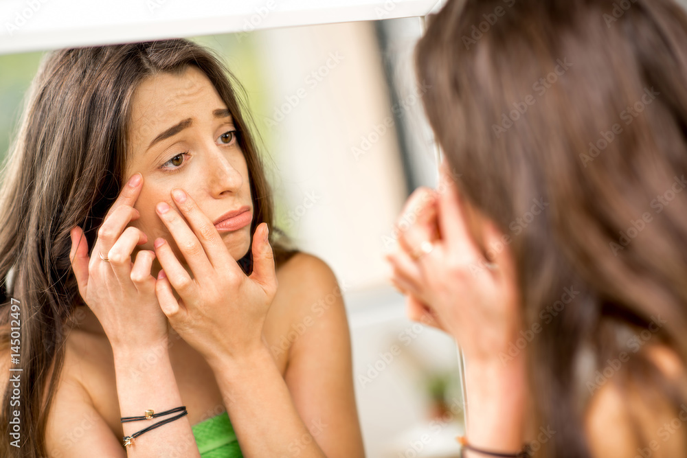 Young sad woman squeezing acne looking at the mirror in the bathroom Young sad woman squeezing acne looking at the mirror in the bathroom