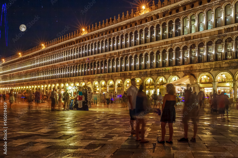 Piazza San Marco, Venice, Italy, illuminated at night with lots of unrecognizable people, colorful sky, stars and full moon