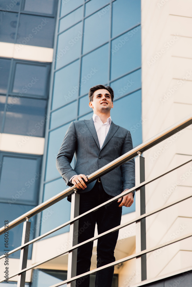 Attractive young confident businessman in classic suit is looking away, leaning on balcony, outside the office building Attractive young confident businessman in classic suit is looking away, leaning on balcony, outside the office building