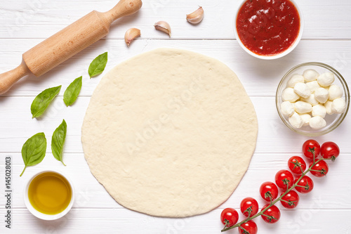 Pizza preparation. Baking ingredients on the kitchen table: rolled dough, mozzarella, tomatoes sauce, basil, olive oil, tomatoes, cheese, spices.
