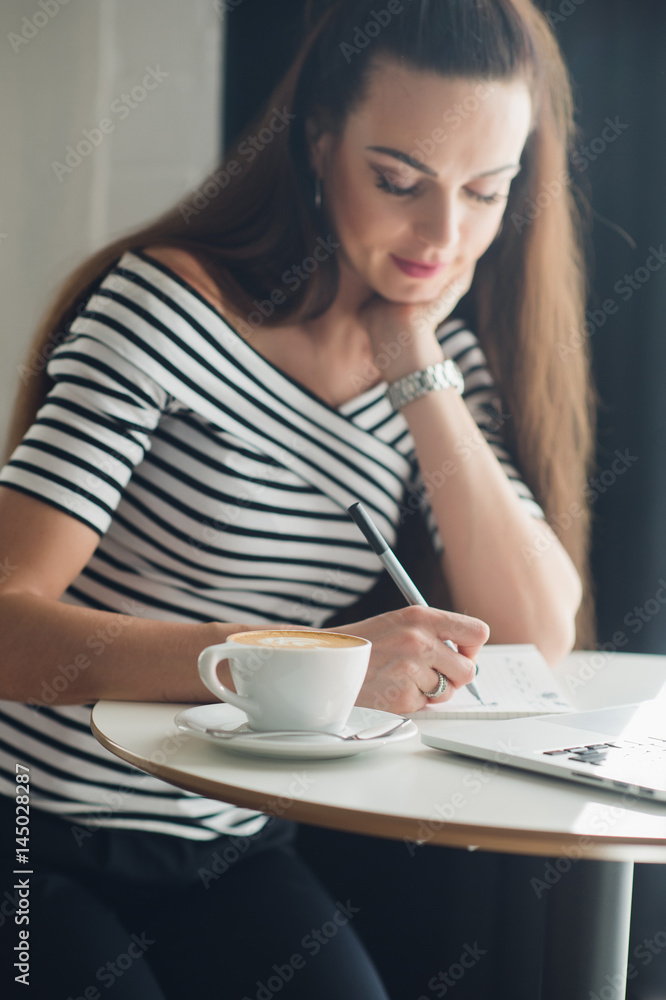 Smiling female writer is working with her new idea at lunch. Attractive adult woman sitting at the table with a cup of coffee and a laptop. Smiling female writer is working with her new idea at lunch. Attractive adult woman sitting at the table with a cup of coffee and a laptop.