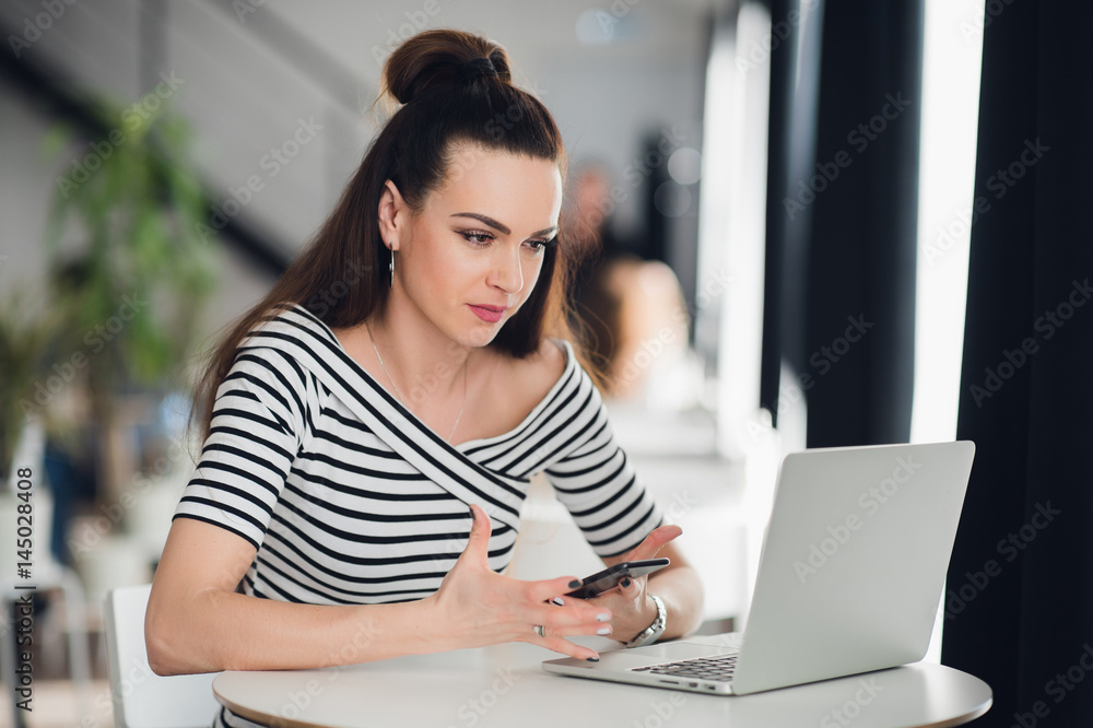 Puzzled woman writer working on a new idea for her book. Adult attractive woman sitting at the table near laptop and holding the phone. Puzzled woman writer working on a new idea for her book. Adult attractive woman sitting at the table near laptop and holding the phone.