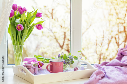 Cozy home concept. Purple fresh tulips in glass vase. Macaroons in glass jar. Cup of hot tea. White tray. Lilac blanket on the windowsill. Sunshine. Coloring and processing photo