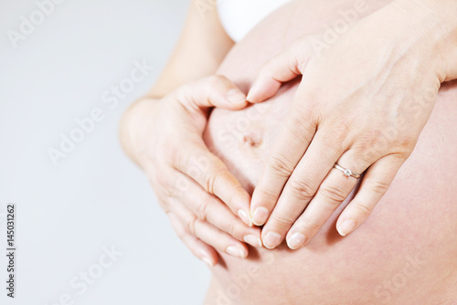 Pregnant woman with shaping heart on her belly with her hands, on grey background, unrecognizable person.