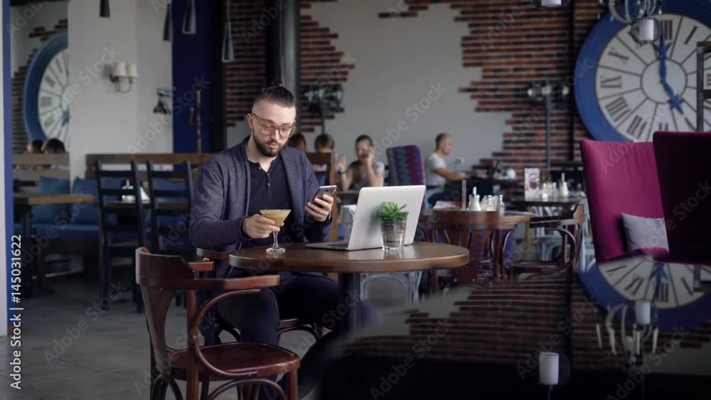 A young attractive hipster in a cafe, with a modern interior. A man is sitting at a table with coffee, using a smartphone, next to his laptop. A young attractive hipster in a cafe, with a modern interior. A man is sitting at a table with coffee, using a smartphone, next to his laptop.