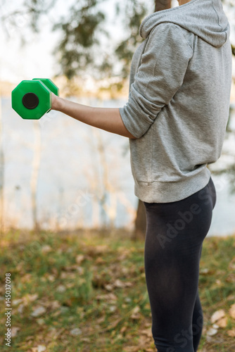 Girl in sports pants and hoodie standing up shakes her bicep hands with green dumbbell