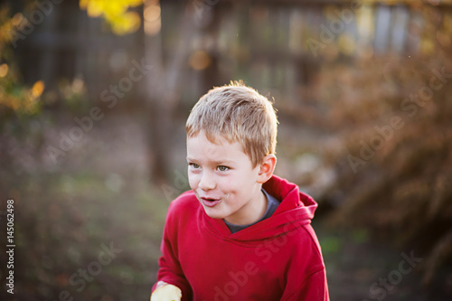 A cheerful boy with an apple rejoices, jumps and dances in summer in nature in the rays of the setting sun. A cheerful child in a red sweater and black pants. A boy eats an apple and grimaces