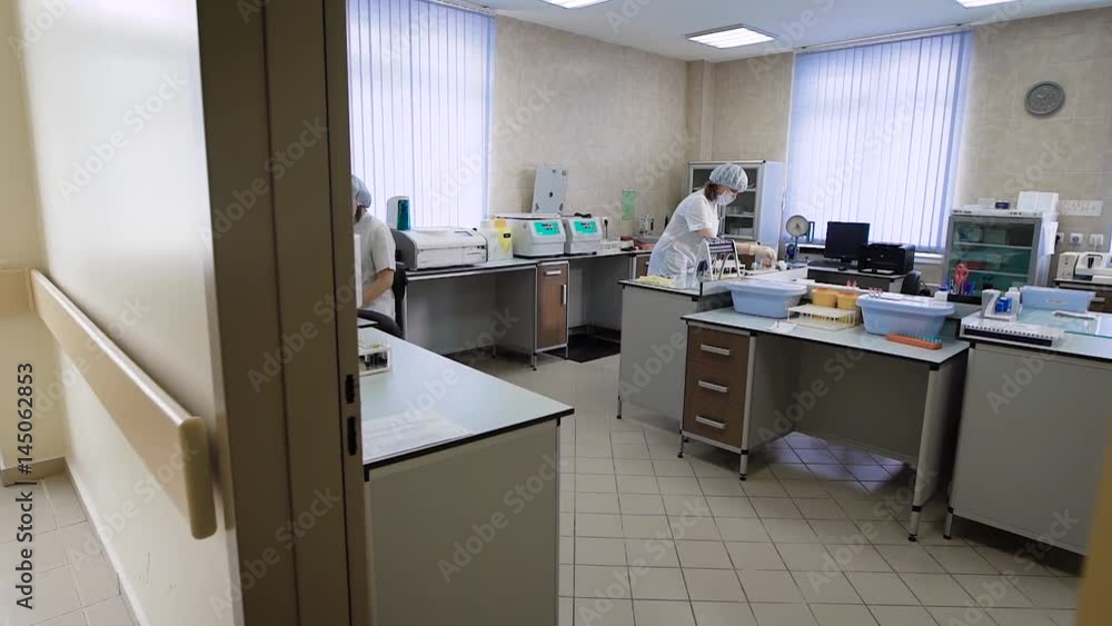 Image of medical laboratory with two female research assistans working inside. Nurse is sitting on the chair at the table. Woman in blue trousers is standing and loading test tubes into centrifuge.