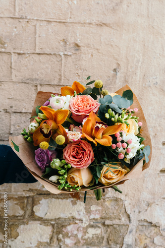 Woman holds colorful  bouquet of different fresh flowers against brick wall. Bunch of orchids, roses, freesia and eucalyptus leaves.