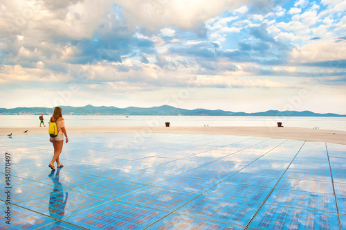 A girl walking on top of Sun Salutation installation made of solar photovoltaic cells near the sea shore against the background of a hill range and dramatic cloudy morning sky. Zadar, Croatia