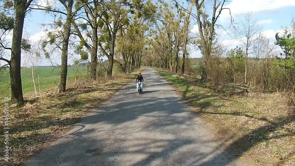 A girl is riding a moped on an alley with trees A girl is riding a moped on an alley with trees
