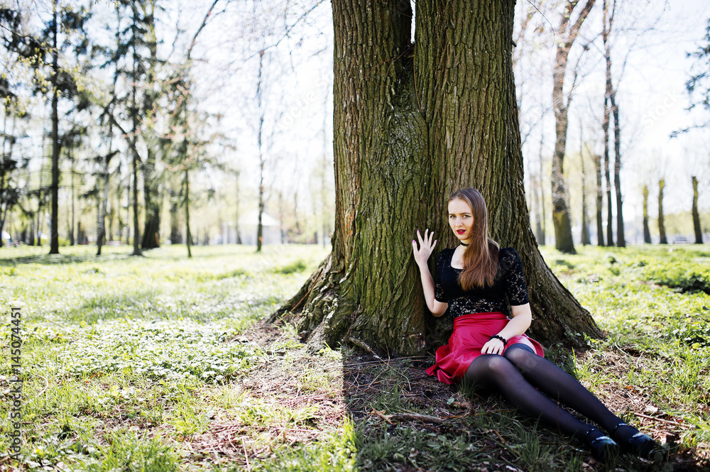 Portrait of girl with bright make up with red lips, black choker necklace on her neck and red leather skirt sitting near tree at park. Portrait of girl with bright make up with red lips, black choker necklace on her neck and red leather skirt sitting near tree at park.