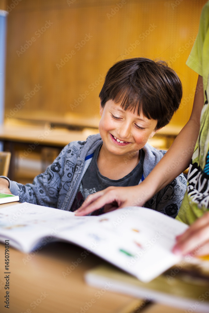 Smiling boy reading book while classmate standing at desk in classroom Smiling boy reading book while classmate standing at desk in classroom