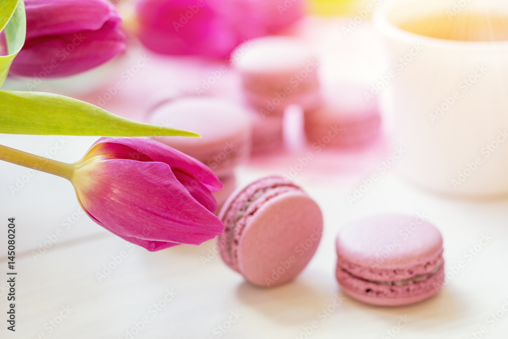 Violet sweet delicious macaroons and fresh tulips on white background. Cup of hot tea. Shallow depth of field. Coloring toned photo. Violet sweet delicious macaroons and fresh tulips on white background. Cup of hot tea. Shallow depth of field. Coloring toned photo.