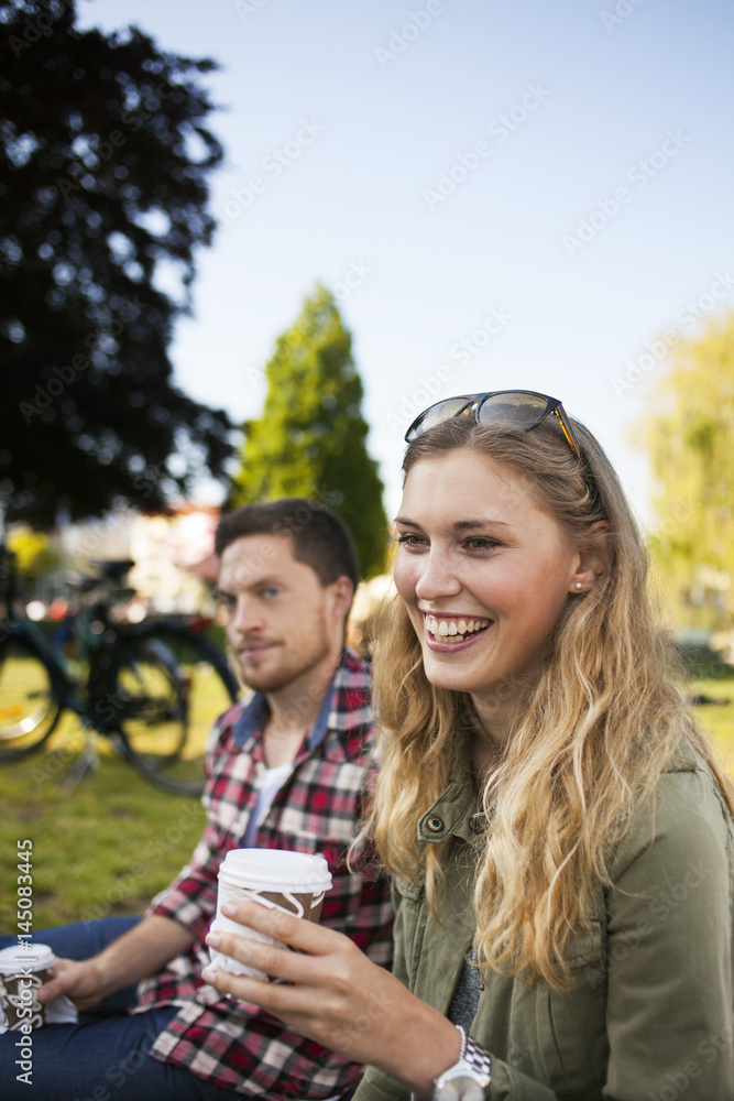 Happy woman holding disposable cup while sitting with friend at park Happy woman holding disposable cup while sitting with friend at park