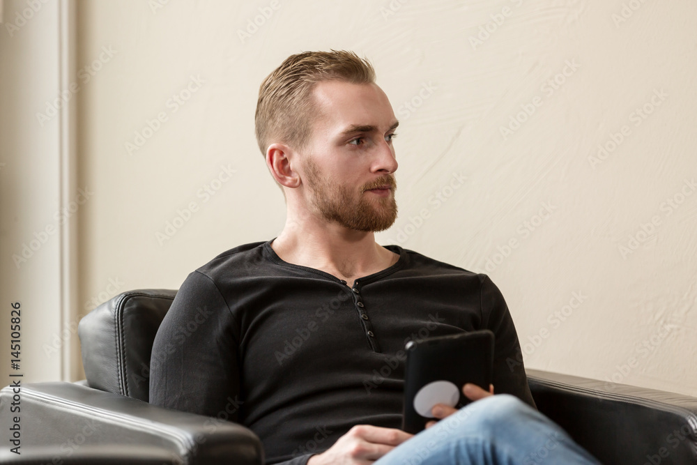 Casual laid back man wearing a black sweater and jeans, sitting down in a lounge chair indoors, holding a digital tablet. Casual laid back man wearing a black sweater and jeans, sitting down in a lounge chair indoors, holding a digital tablet.