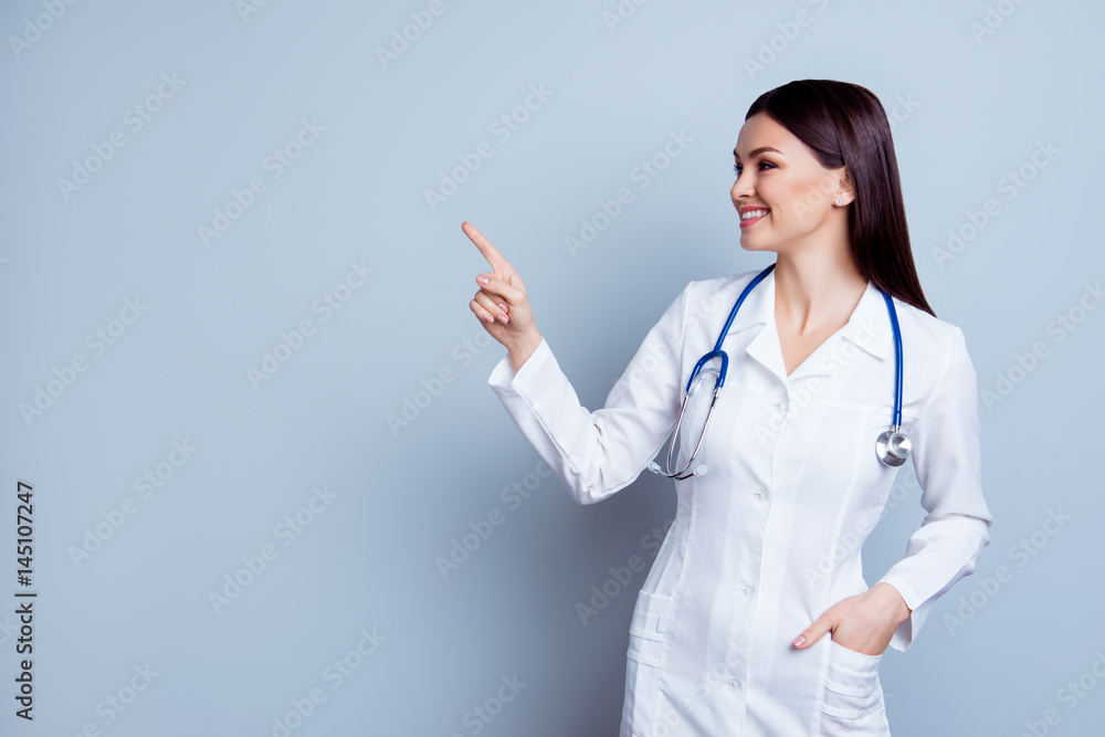 Horizontal portrait of young pretty smiling doc depicted on gray background holding hand in a pocket and pointing on copy space Horizontal portrait of young pretty smiling doc depicted on gray background holding hand in a pocket and pointing on copy space