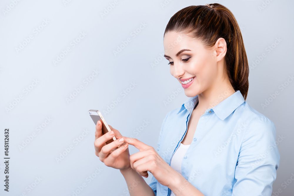 Close up photo of attractive smiling girl standing against gray background and typing sms on her smartphone Close up photo of attractive smiling girl standing against gray background and typing sms on her smartphone