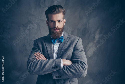 Portrait of serious young bearded man with mustache in a suit stands on a gray background with  folded his hands and fixedly looks