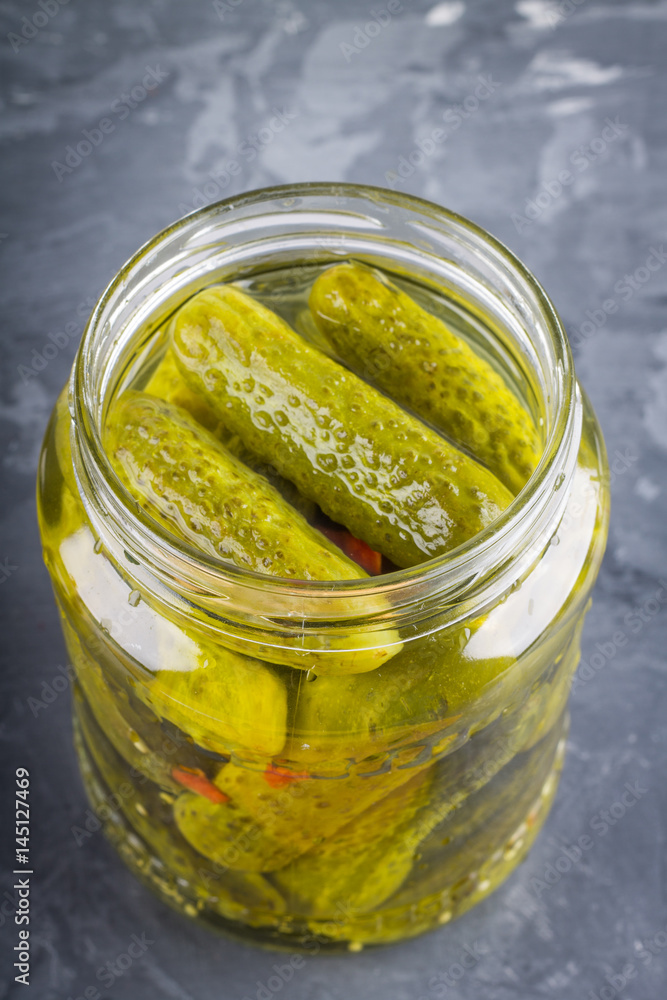 Pickled gherkins or cucumbers in glass jar on a gray background