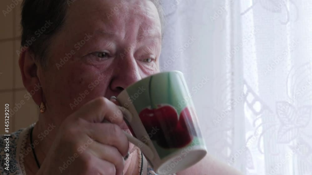 An elderly woman holds a cup of hot drink and drinks. He sits near the window and has breakfast. Simple home clothes. An elderly woman holds a cup of hot drink and drinks. He sits near the window and has breakfast. Simple home clothes.
