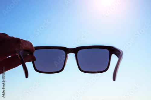 Male hand holding sunglasses in front of sun, blue sky on background