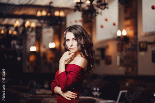 Beautiful European girl in a long cocktail evening dress of red coral with curly hair is standing in the restaurant. Concept of the make-up artist s work.